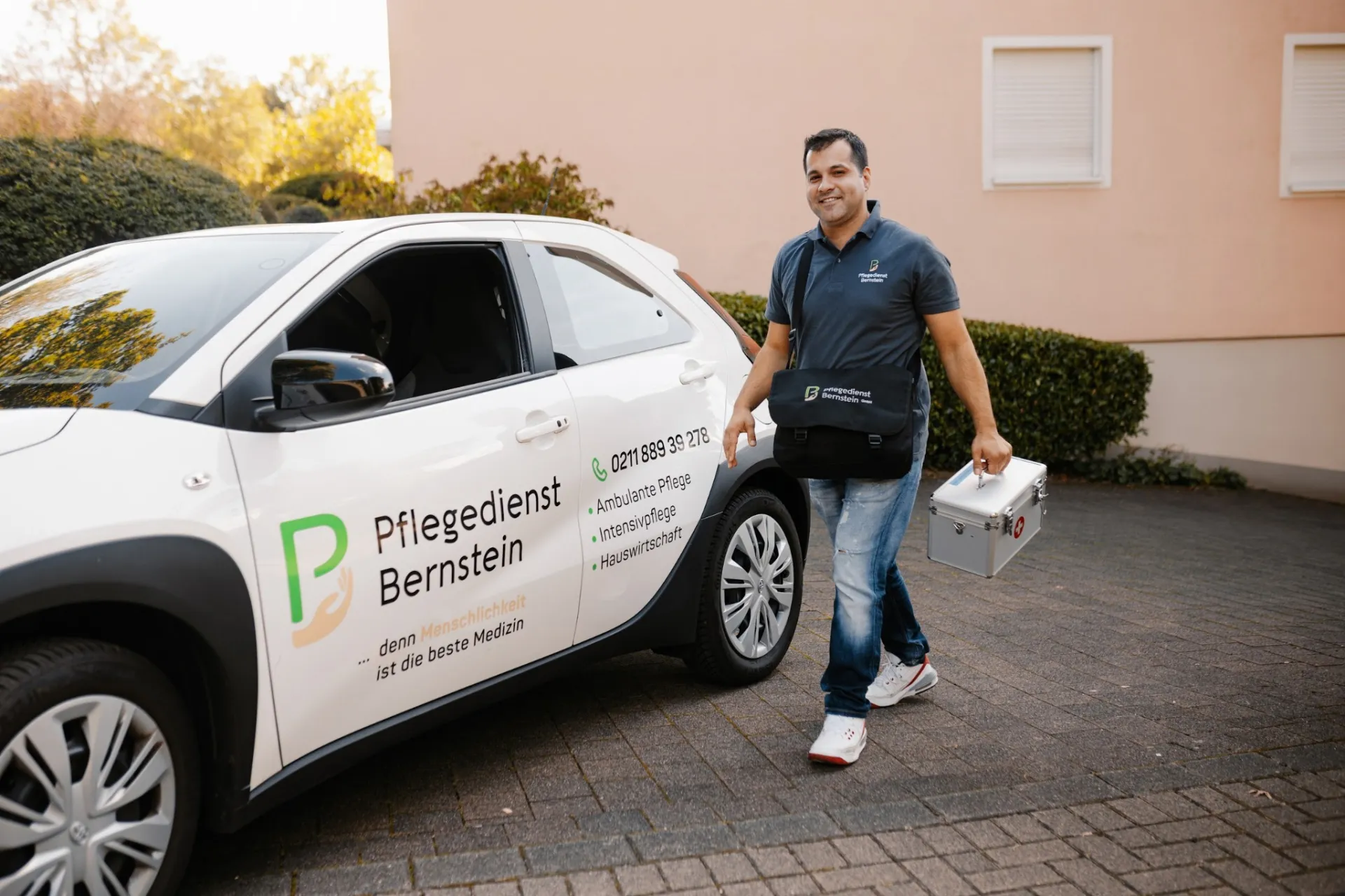 Ein Pfleger von Bernstein in dunklem Dienstpoloshirt geht mit Umhängetasche und Pflegekoffer lächelnd an einem beschrifteten Firmenwagen des Pflegedienstes vorbei.